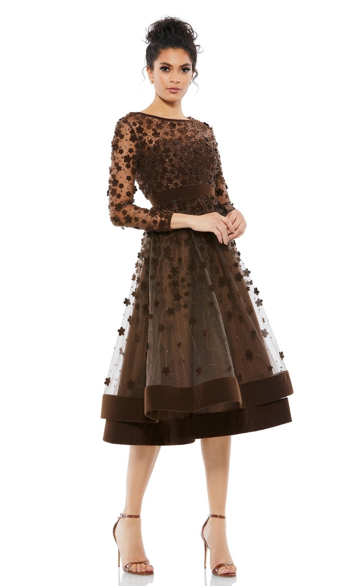 A woman wears the Mac Duggal 67007 knee-length wedding-guest dress with floral embellishments, sheer long sleeves, appliqués, and a tulle overlay, paired with strappy high-heeled sandals.